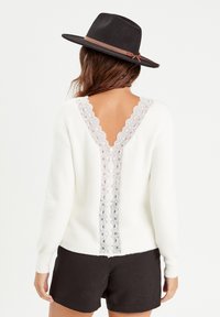 Pull blanc avec un détail en dentelle dans le dos en V, présentant une texture douce et des manches longues, associé à un short noir et un chapeau noir.