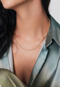 Złoty skręcony naszyjnik leży na skórze, dopełniając jedwabistą zielono-niebieską bluzkę o gładkiej fakturze i miękkim układzie.