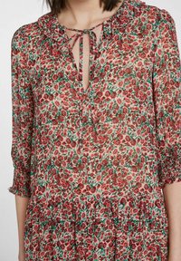 Blouse à imprimé floral en tissu transparent, avec un col à nouer, des manches volantes et un design à niveaux avec des motifs de fleurs rouges et vertes.