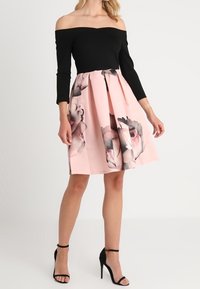 Svart off-shoulder topp i kombination med en rosa kjol med stora blommönster; mjukt tyg, plisserad design och svarta högklackade skor.