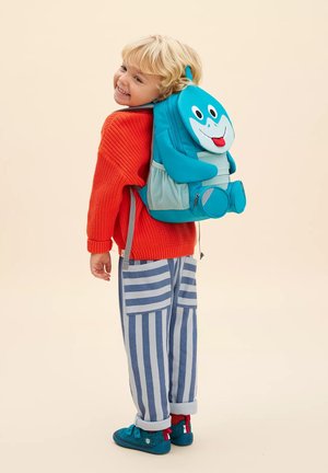 Junges Kind in orangefarbenem Pullover und gestreifter Hose lächelt, während es einen blauen verspielten Haifisch-Rucksack auf beigem Hintergrund trägt.