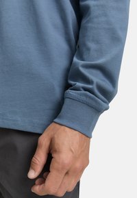 Lange mouwen, blauwe katoenen shirt met een ronde kraag en smalle manchetten. Kenmerkt een soepele textuur en een afgeronde zoom. Hand zichtbaar aan de onderkant.