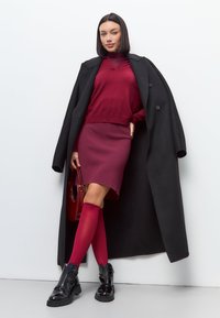 Rode trui en rok, zwarte jas, glanzende zwarte laarzen en rode kniekousen. Gladde stoffen met gestructureerde vormen en minimale details.