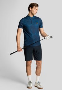 Polo blu navy con texture a motivo, maniche corte e un logo, abbinato a pantaloni corti scuri e scarpe. Regge un bastone da golf.
