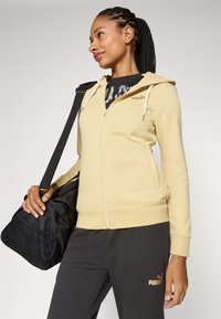 Puma Kapuzenpullover mit Reißverschluss aus weichem, gelbem Stoff, mit Frontreißverschluss, Kordelzugkapuze und Seitentaschen, kombiniert mit dunkelgrauen Sweatpants und einer schwarzen Tasche.
