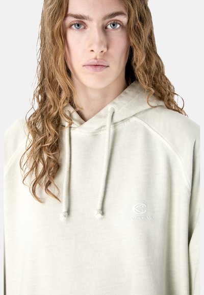 Jeune femme aux cheveux blonds ondulés portant un sweat à capuche beige clair avec des cordons de serrage et un petit logo Camperlab sur la poitrine.