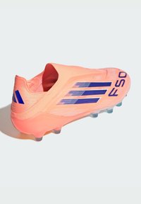 Bota de fútbol de color coral brillante con rayas azules de Adidas y la letra "FSO", con un diseño sin cordones y suela con tacos para jugar en terrenos firmes.