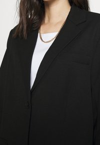 Musta bleiseri, jossa on teksturoitu pinta, yksi nappi kiinnityksessä ja rintatasku, päällä valkoinen ribbitekstiilistä valmistettu toppi ja kullanvärinen ketju.