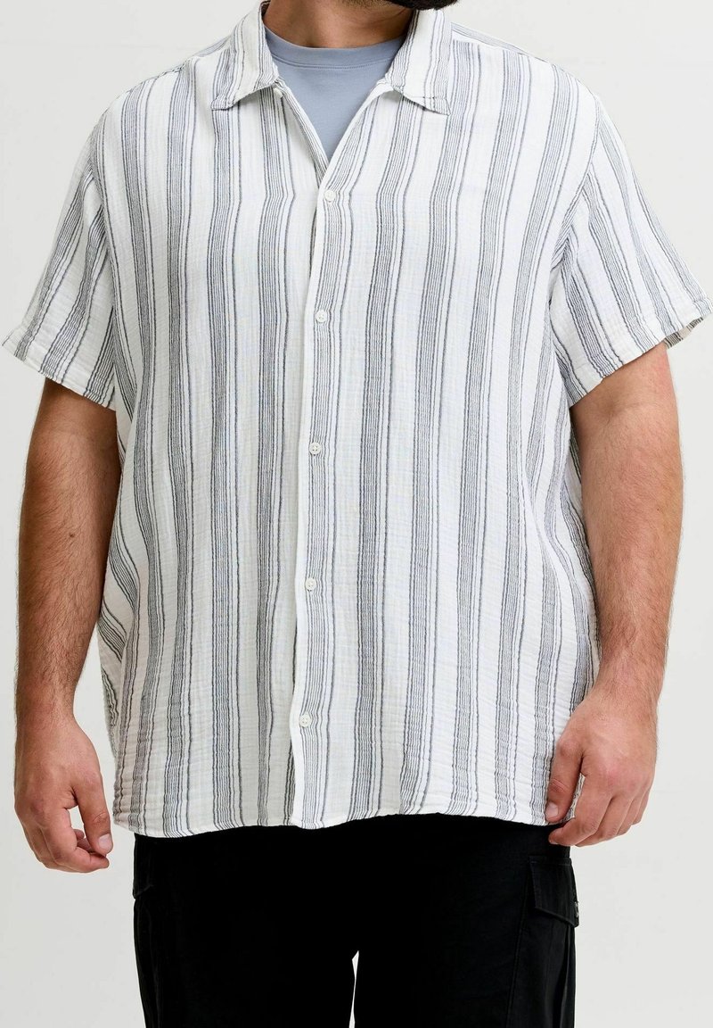 Homme portant une chemise blanche à manches courtes à boutons avec des rayures noires verticales sur un t-shirt gris et un pantalon noir sur un fond uni.