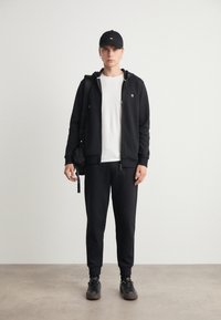 Felpa nera con cappuccio, t-shirt bianca sotto, pantaloni jogger neri e sneakers neri. Gli accessori includono un cappellino nero e una borsa piccola.