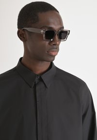 Klare schwarz gerahmte Sonnenbrille mit dunklen Gläsern, getragen zu einem schwarzen Hemd mit Kragen. Glattes, modernes Design mit subtilen Strukturdetails.