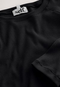 Zwart geribd textiel met een ronde halslijn, voorzien van een wit label met zwarte tekst "self." in een vloeiende typografie. Zachte textuur, casual ontwerp.