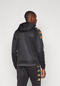 Black hoodie made of smooth fabric, featuring a contrasting gold logo on the shoulders. Designed with a hood and fitted silhouette.