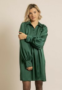 Robe en satin vert avec un col, des manches longues bouffantes et une coupe ample. Texture lisse et longueur genou, avec des plis discrets.