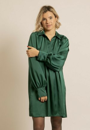 Robe en satin vert avec un col, des manches longues bouffantes et une coupe ample. Texture lisse et longueur genou, avec des plis discrets.