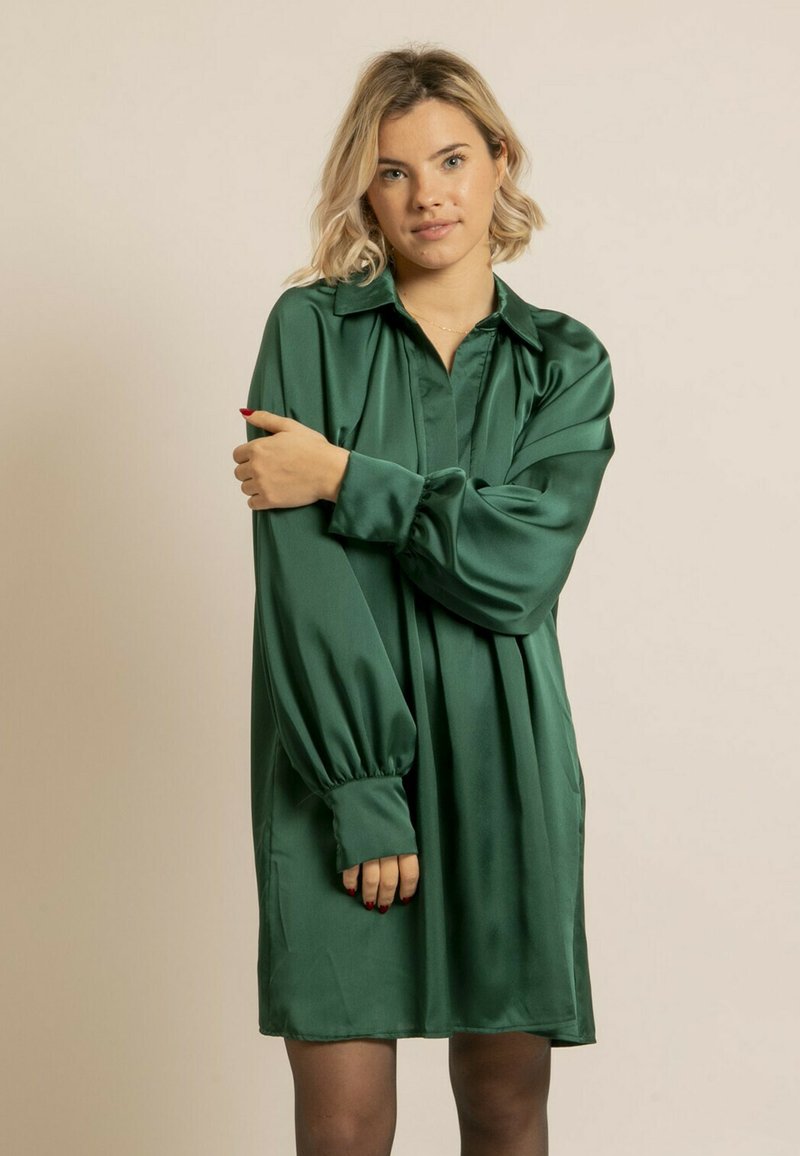Robe en satin vert avec un col, des manches longues bouffantes et une coupe ample. Texture lisse et longueur genou, avec des plis discrets.