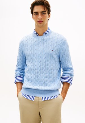 Jaunas vyras dėvi šviesiai mėlyną pynimo megztinį ant mėlynai baltos dryžuotos marškinių, smėlio spalvos kelnes, rankos kišenėse.