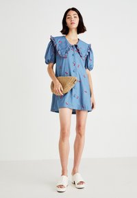 Džínové šaty s nataženými rukávy a velkým volánkovým límcem; zdobí je výšivka třešní a přední knoflíky, doplněné tkanou kabelkou.