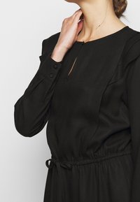 Schwarzes Kleid mit rundem Ausschnitt, Schlüsselloch-Detail, Rüschen an den Schultern, Knopfmanschetten und einem Taillengürtel für eine verstellbare Passform. Weicher Strukturstoff.