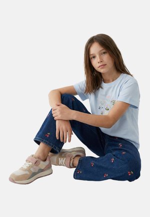 Jeune fille aux longs cheveux bruns assise en tailleur portant un t-shirt bleu clair à motif floral, un jean foncé brodé de cerises et des baskets beiges.