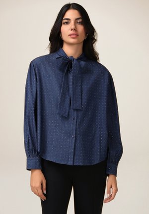Blouse bleu marine avec un col à nœud, manches longues et boutonnage à l'avant. Le tissu présente de petits pois métalliques, uniformément espacés.