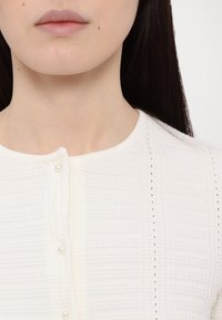 Fehér textúrált kötött felső, lekerekített nyakkivágással, gombos elejével, gyöngyszerű gombokkal és függőleges vonal részletezéssel.