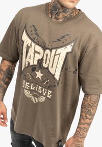 Barna, oversized póló, ami egy megviselt "TAPOUT" grafikát tartalmaz fehér színben, csillag és szárny díszítéssel. Puha pamutanyagból készült.