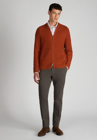 Joven con un suéter de punto naranja óxido con cremallera, camiseta de rayas, pantalones gris oscuro y botas de ante marrón, de pie contra un fondo liso.