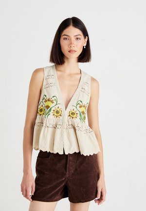 Top sans manches crème avec broderie florale en jaune et vert, accents en dentelle et ourlet à volants ; associé à un short marron foncé.