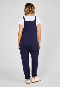 Marineblauer Overall mit verstellbaren Trägern, bequemer Passform und umgeschlagenen Knöcheln, kombiniert mit einem weißen T-Shirt und Sneakers.