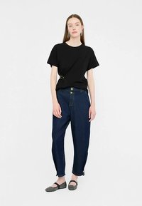 Schwarzes T-Shirt mit kurzen Ärmeln und Schnallendetail, kombiniert mit hoch taillierten dunkelblauen Jeanshosen mit goldenen Knöpfen und konisch zulaufenden Beinen.