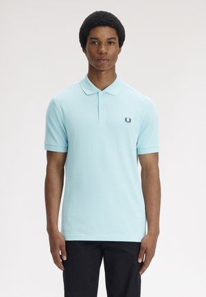 Jeune homme aux cheveux afro courts portant un polo à manches courtes bleu clair et un pantalon noir, debout devant un fond clair uni.