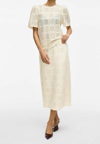 Kvinna i krämfärgad virkad topp och matchande midikjol, tillsammans med mörkbruna spetsiga klackskor.