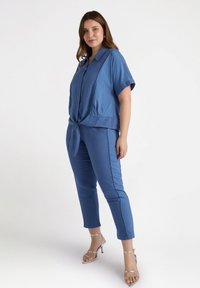 Blå kortärmad skjorta med knäppning och knytning i midjan, tillsammans med blå figurnära byxor. Modellen bär silverfärgade sandaler med remmar. Slät tygtextur.