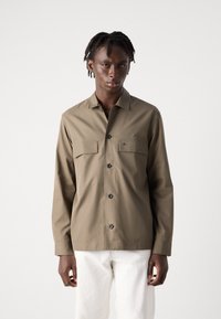 Brown button-up shirt made of smooth fabric, featuring two chest pockets and a collar. Worn with white pants, shown in a neutral setting.