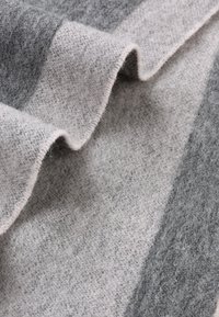 Couverture en laine avec des rayures grises claires et foncées alternées, texture douce et bord replié révélant un motif de tissage subtil.