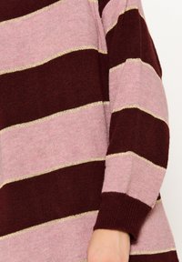 Maglione lavorato a maglia con ampie righe bordeaux e rosa, arricchito da dettagli in filo dorato. Primo piano della manica che mostra la morbidezza del tessuto e il polsino a coste.