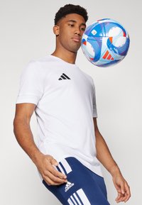 Camisa deportiva blanca con logo negro, combinada con pantalones azules que tienen rayas blancas, mientras un balón de fútbol colorido flota cerca del torso.