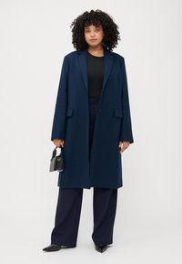 Una persona con capelli ricci indossa un cappotto blu navy sopra una maglietta nera, tenendo una piccola borsa nera. Si trova di fronte a uno sfondo bianco uniforme.