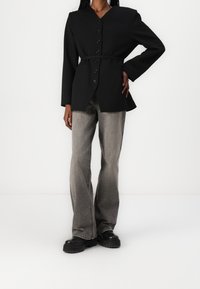 Černý saku s knoflíky a páskem, přizpůsobený střih, dlouhé rukávy a kapsy. V kombinaci s rozšířenými šedými d�žíny a robustními černými botami.