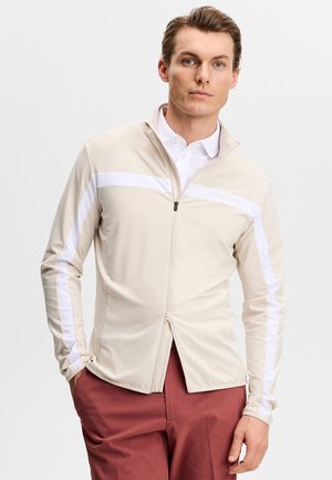 Homme portant une veste beige à fermeture éclair avec des bandes blanches sur les manches et la poitrine, associée à un pantalon couleur rouille, debout devant un fond uni.