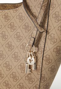 Bruine tote bag met een patroon en een gestructureerde afwerking, met een sleutelhanger met een sleutel en twee decoratieve charms, en contrasterende donkere rand.