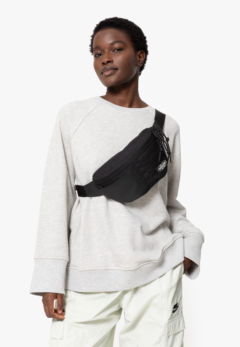 Marsupio nero con doppie tasche con zip e cintura regolabile. Il modello indossa un dolcevita grigio chiaro e pantaloni cargo chiari.