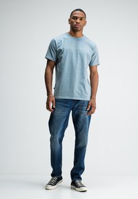 Jonge man staat in een lichtblauwe graphic t-shirt, blauwe jeans en zwarte high-top sneakers tegen een effen witte achtergrond.