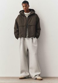 Donkerbruine gewatteerde jas met capuchon en voorzakken, gecombineerd met grijze wijdere sweatpants en beige sneakers. Witte en gestreepte shirt eronder.