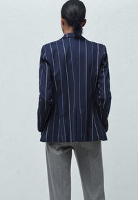 Granatowa marynarka w prążki, dopasowana, z pojedynczym rozcięciem i usztywnionymi ramionami, zestawiona z szarymi spodniami. Gładka tekstura materiału.