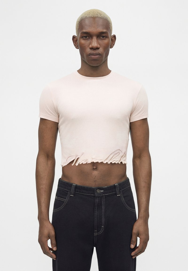 Modèle masculin aux cheveux blonds courts portant un t-shirt court rose clair et un jean denim foncé sur un fond uni.