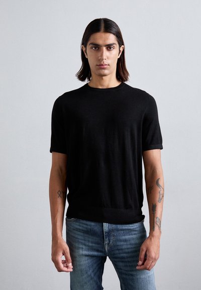 Schwarzes kurzärmliges Strick-T-Shirt mit Rundhalsausschnitt. Das Modell kombiniert es mit hellblauen Jeans. Weiche Textur, lässige Passform.