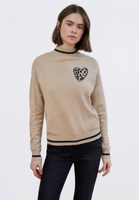 Maglione beige con collo alto, caratterizzato da un dettaglio a cuore nero con stampa leopardata sul petto. Dettagli neri a coste su polsini e orlo.