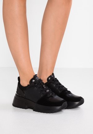 Odzież na nogi pokazująca czarne sneakersy na platformie z mieszanymi materiałami i sznurowadłami na jasnej podłodze i tle.